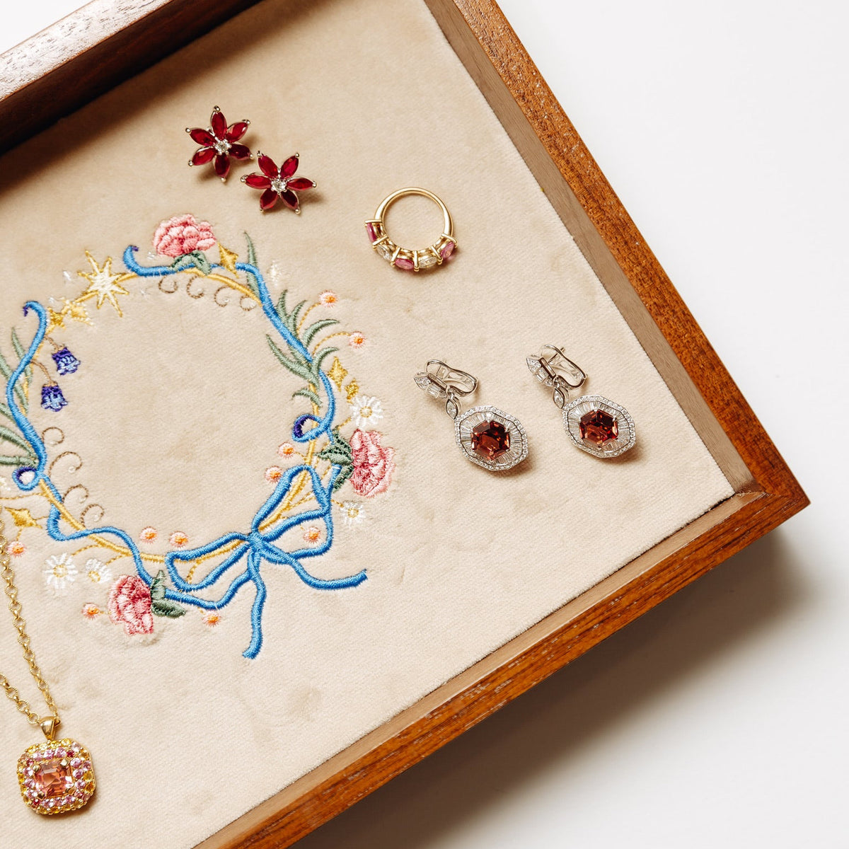 L’HERITAGE Fine Jewelry gemstone collection displayed in a vintage wooden tray with embroidered floral details, featuring ruby flower earrings, a gold gemstone ring, sparkling garnet earrings, and a pink gemstone pendant necklace.