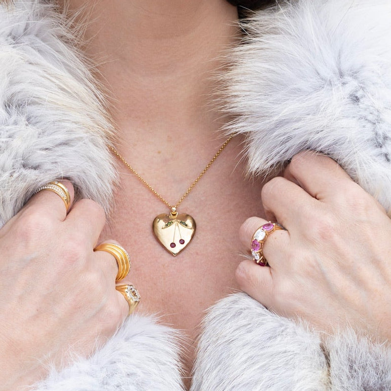 A close-up lifestyle shot of a person wearing a gold heart pendant necklace with a small red cherry detail, styled with a white fur jacket and gold rings.