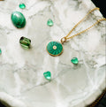 A gold pendant with a green swirled stone and a central diamond on a chain, displayed on a white marble surface with loose green gemstones nearby.