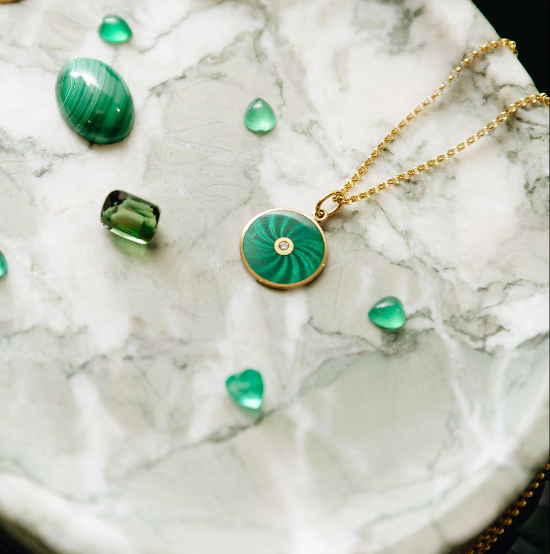 A gold pendant with a green swirled stone and a central diamond on a chain, displayed on a white marble surface with loose green gemstones nearby.