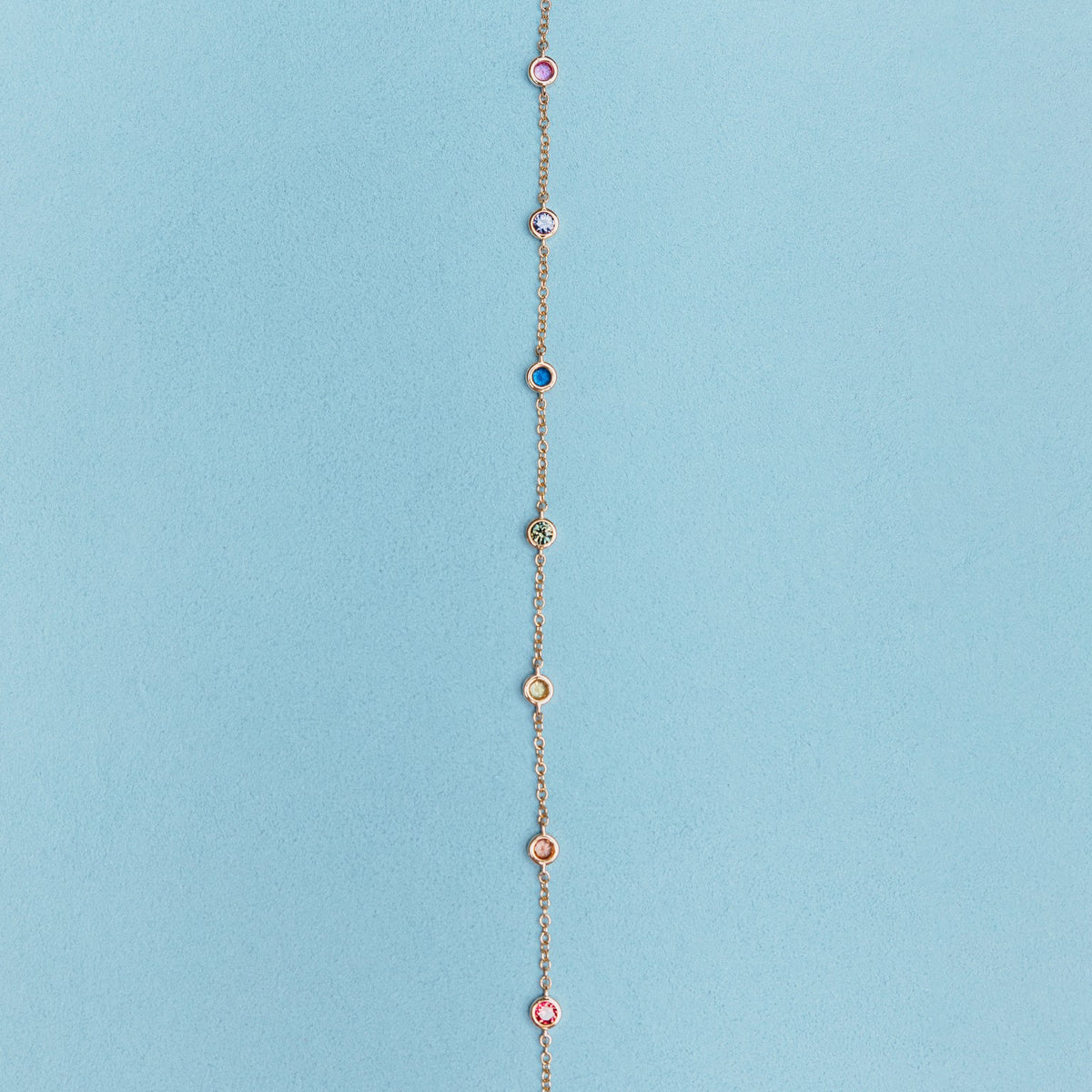 A delicate gold chain bracelet featuring nine evenly spaced, small circular enamel medallions in various colors on a solid light blue background.