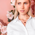Woman wearing a red initial enamel guilloche pendant and a white shirt with a necklace against a floral background. Initial Guilloché Pendant. Crafted in 18k yellow gold with translucent guilloché enamel. 15mm diameter. Jewelry Dallas.