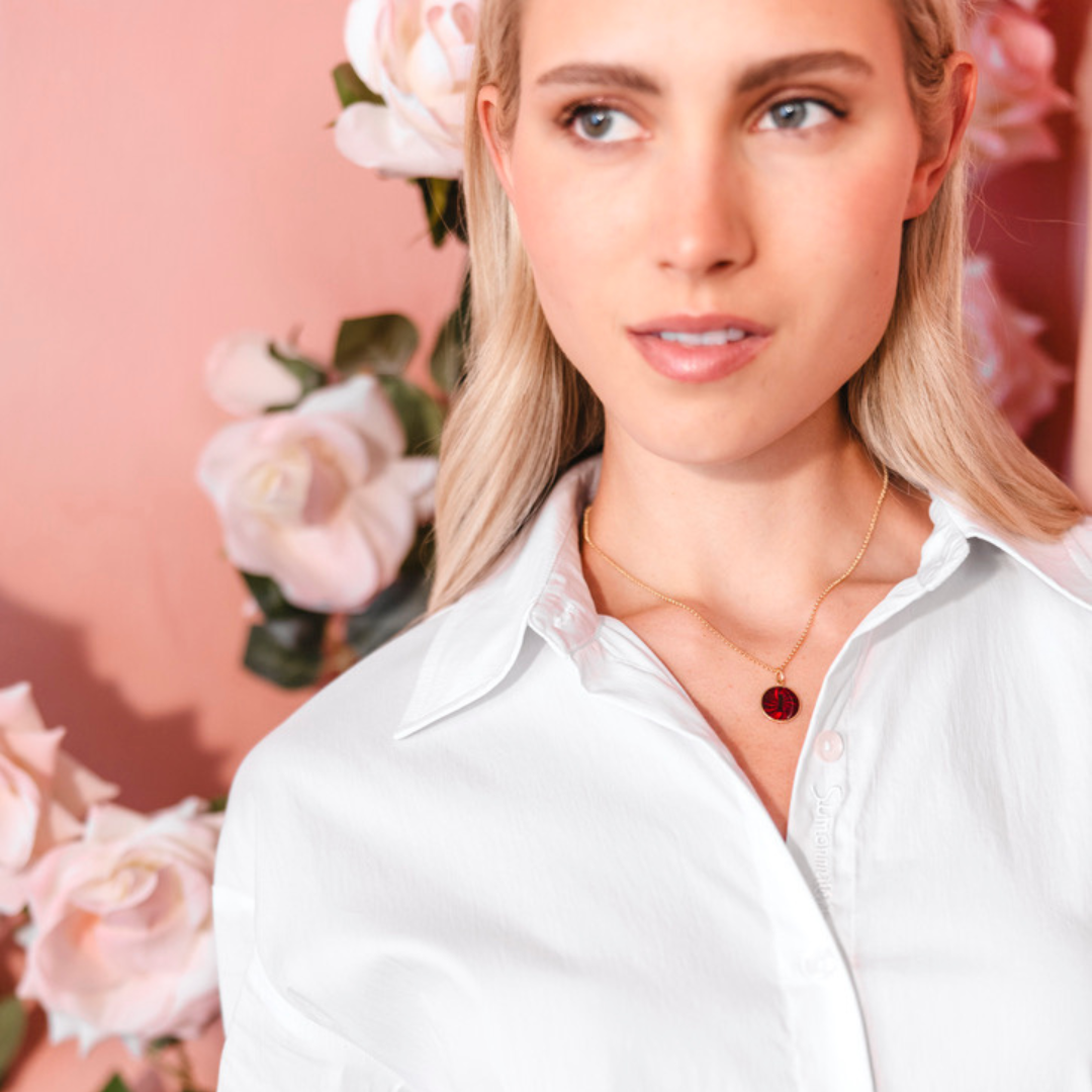 Woman wearing a red initial enamel guilloche pendant and a white shirt with a necklace against a floral background. Initial Guilloché Pendant. Crafted in 18k yellow gold with translucent guilloché enamel. 15mm diameter. Jewelry Dallas.