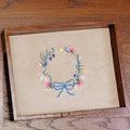 An embroidered floral wreath design on beige fabric, framed in a wooden jewelry tray, photographed on a wooden tabletop.