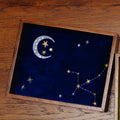 A wooden jewelry tray with a dark blue velvet insert featuring embroidered celestial motifs, including a silver crescent moon, stars, and constellations, shown on a wooden tabletop.