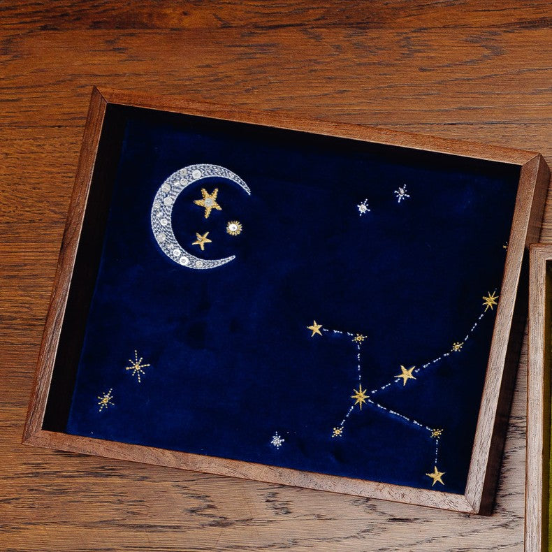 A wooden jewelry tray with a dark blue velvet insert featuring embroidered celestial motifs, including a silver crescent moon, stars, and constellations, shown on a wooden tabletop.