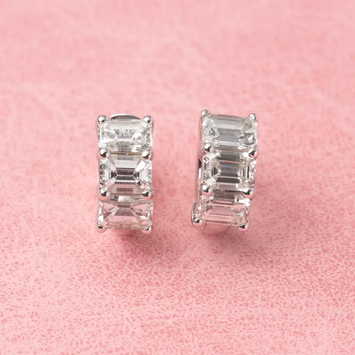 Diamond huggie earrings with baguette cut stones on a pink background. 3-Stone Emerald Huggies. Solid gold and natural diamonds. Jewelry Dallas.