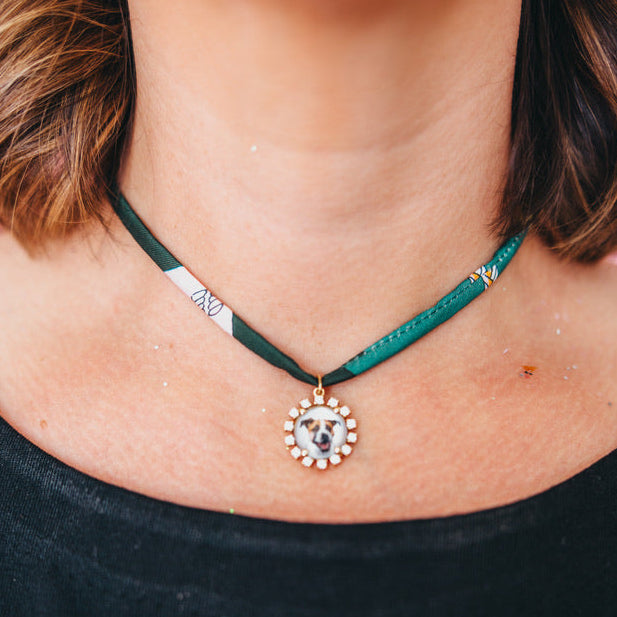 A close-up of a person's neck and chest wearing a delicate gold chain necklace with a custom circular pendant featuring a dog portrait surrounded by small clear stones.