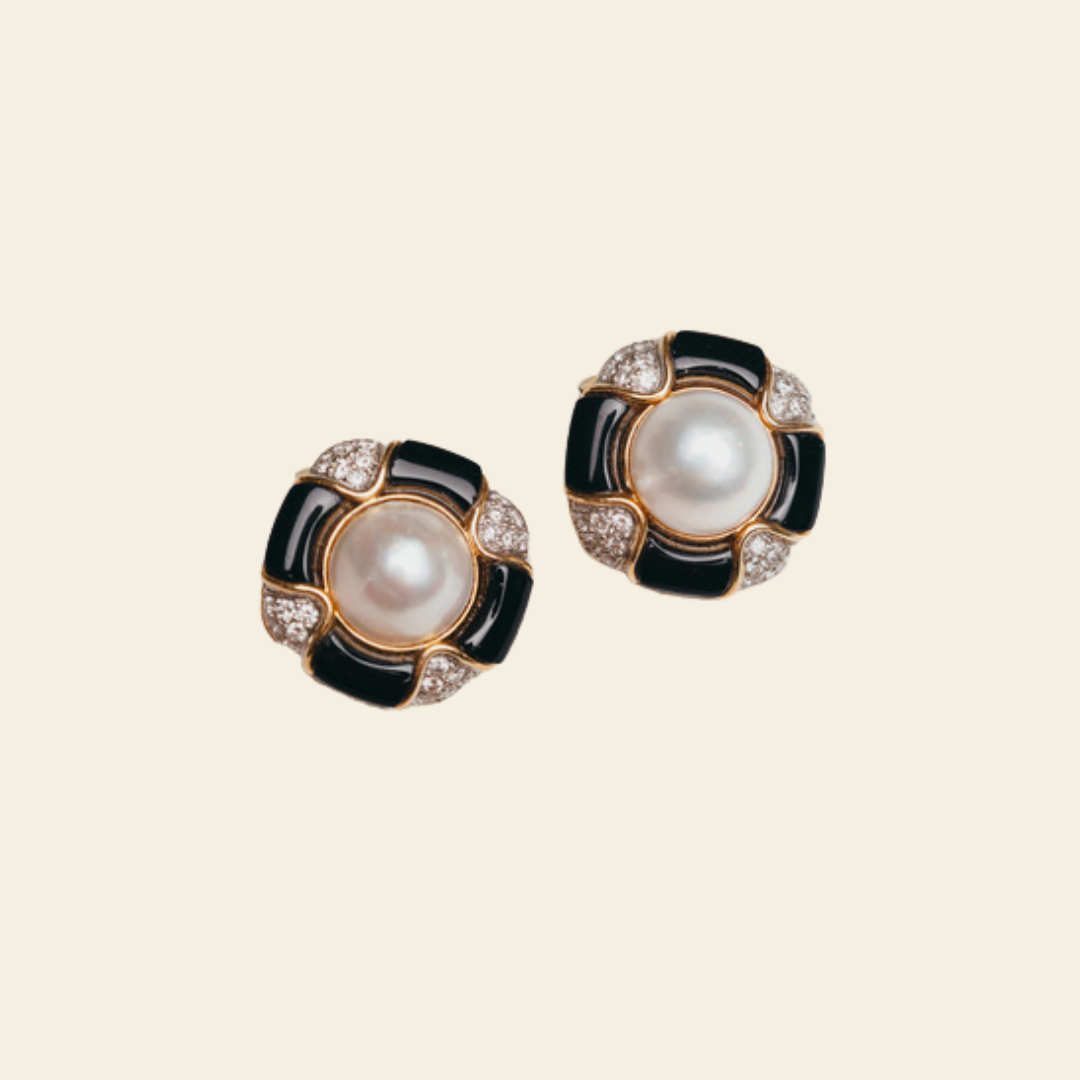 A pair of stud earrings featuring a central white pearl surrounded by black enamel and small clear stones in a gold-tone setting, displayed on a solid cream background.