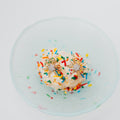 A top-down view of an ice cream on a pale blue glass plate, topped with colorful sprinkles and two gold starburst-shaped earrings with clear rhinestone centers.