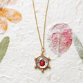 A gold chain necklace with a round pendant featuring a red and black ladybug illustration under clear cover, displayed on a textured off-white background with subtle pressed floral elements.