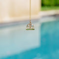 A gold pendant necklace featuring a miniature sailboat with a clear glass dome and green gemstone accents, suspended against a blurred background of a swimming pool and outdoor setting.