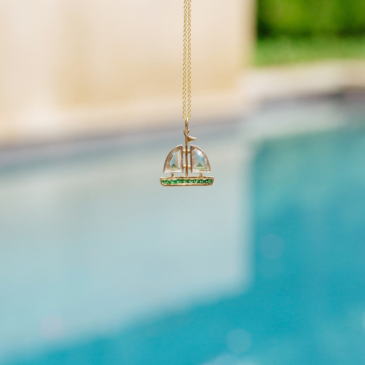 A gold pendant necklace featuring a miniature sailboat with a clear glass dome and green gemstone accents, suspended against a blurred background of a swimming pool and outdoor setting.