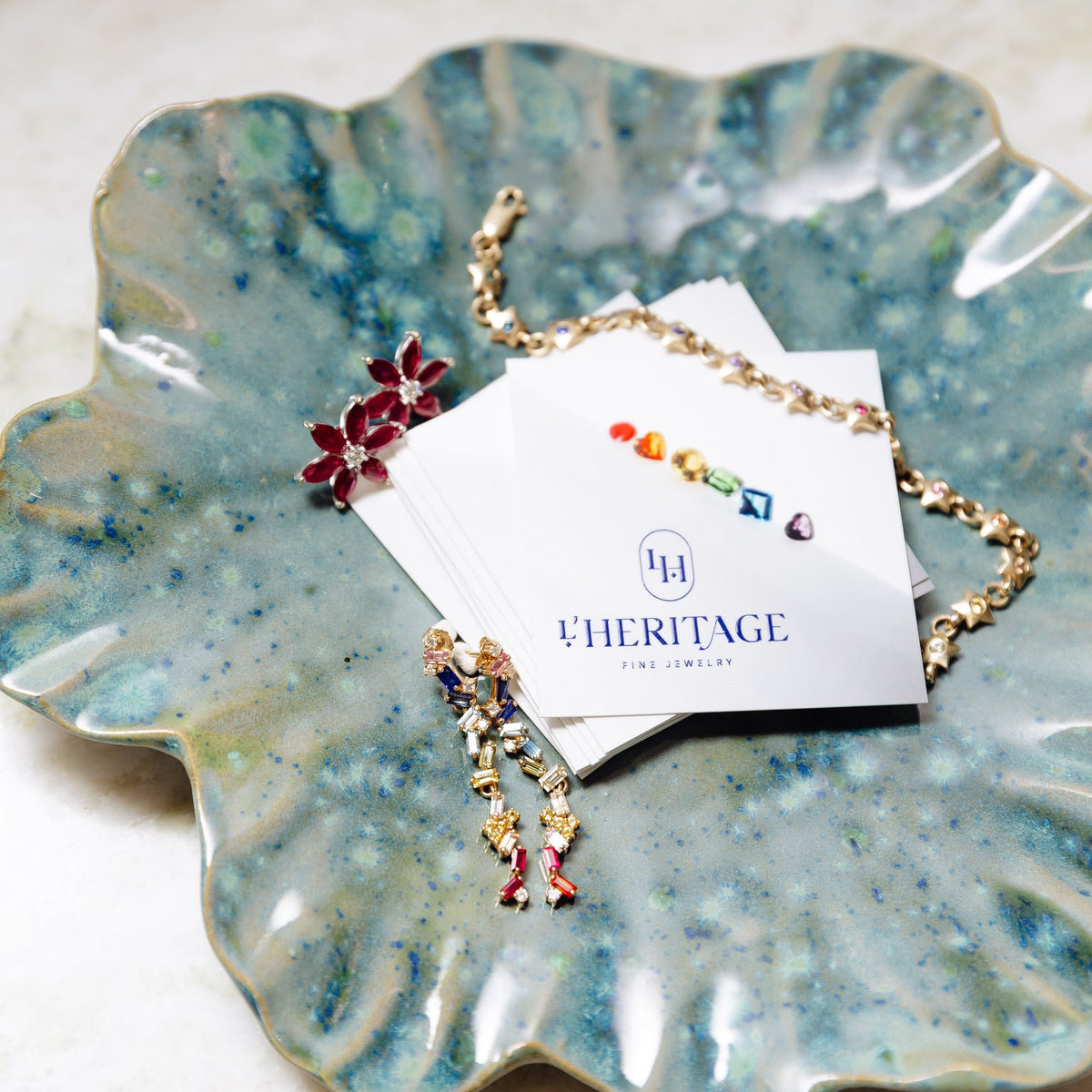 L’HERITAGE Fine Jewelry colorful gemstone jewelry displayed on a handcrafted blue ceramic dish, featuring gold necklaces, ruby flower earrings, and vibrant multicolored gemstone pieces alongside branded jewelry cards.
