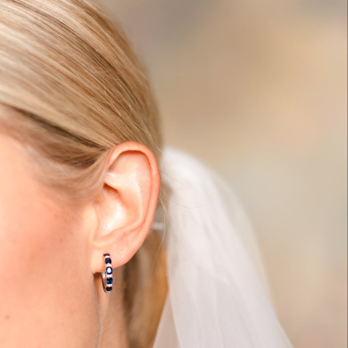 A close-up side profile of a blonde bride wearing a small silver huggie hoop earring with blue sapphires and diamonds, along with a white bridal veil against a soft, blurred background.