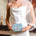 A woman in a white satin wedding dress holds a light blue embroidered travel jewelry box with floral and fairy details. She is wearing a sheer white veil and rings, standing in a softly lit indoor setting.