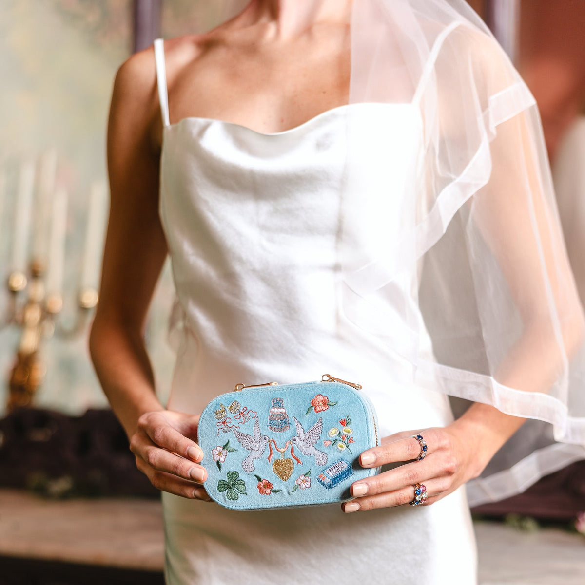 A woman in a white satin wedding dress holds a light blue embroidered travel jewelry box with floral and fairy details. She is wearing a sheer white veil and rings, standing in a softly lit indoor setting.