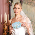 A woman in a white wedding dress and veil holds a light blue embroidered travel jewelry box in a vintage-style room with pink walls, floral wallpaper, and a mirror.