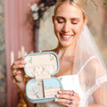 A smiling bride in a white wedding dress and veil holds open a small light blue travel jewelry box with cream lining and floral embroidery. The bride is wearing rings, and the background is a softly blurred indoor setting with warm tones.