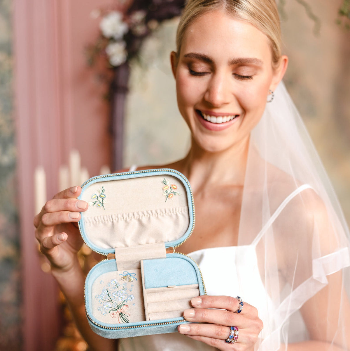 A smiling bride in a white wedding dress and veil holds open a small light blue travel jewelry box with cream lining and floral embroidery. The bride is wearing rings, and the background is a softly blurred indoor setting with warm tones.