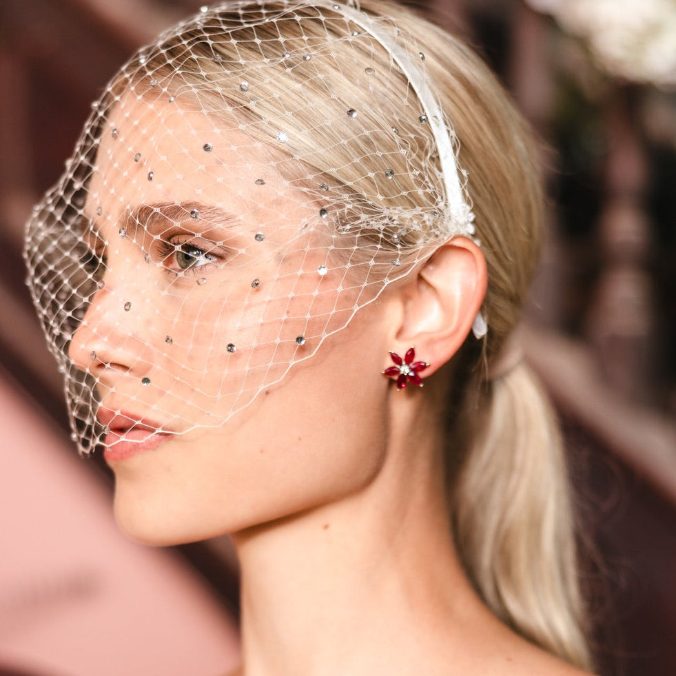 A close-up side profile of a blonde woman wearing a sheer white netted head veil with small rhinestone accents and a matching white headband, paired with a red flower-shaped earring.