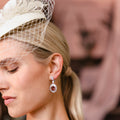 A close-up of a woman wearing a Pink Tourmaline Hexagon Drop Earring with Diamonds, paired with a cream fascinator hat.