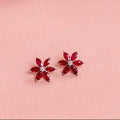 A pair of silver flower-shaped stud earrings with deep red marquise-cut gemstones and a small clear center stone, displayed on a soft pink textured surface.