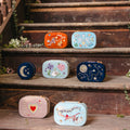 Six embroidered travel jewelry boxes are arranged on rustic wooden stairs decorated with flowers and a draped pink fabric. The embroidered travel jewelry boxes feature various whimsical designs including space-themed patterns, a moon, a heart, and floral motifs in pastel and bright colors.