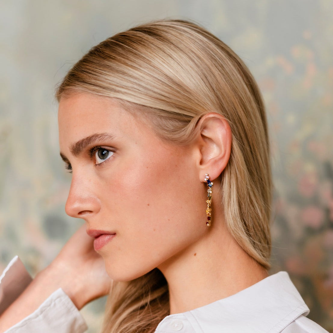 A blonde woman in a white button-up shirt wearing a long, dangling gold earring, shown in profile against a soft, painterly floral background.
