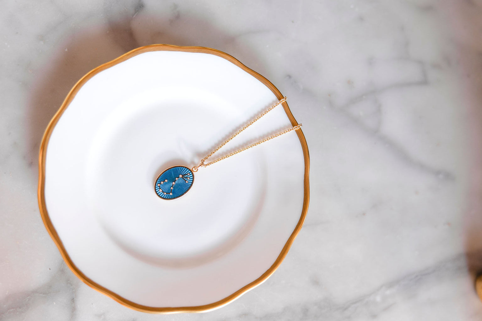 Necklace with a blue enamel guilloche pendant on a white plate with gold rim against a marble backgroundslider_item_h7dLgN