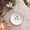Three small stud earrings in red, green, and blue are arranged in a small white ceramic dish with a gold rim, styled on a marble surface with flowers and decorative objects around it.