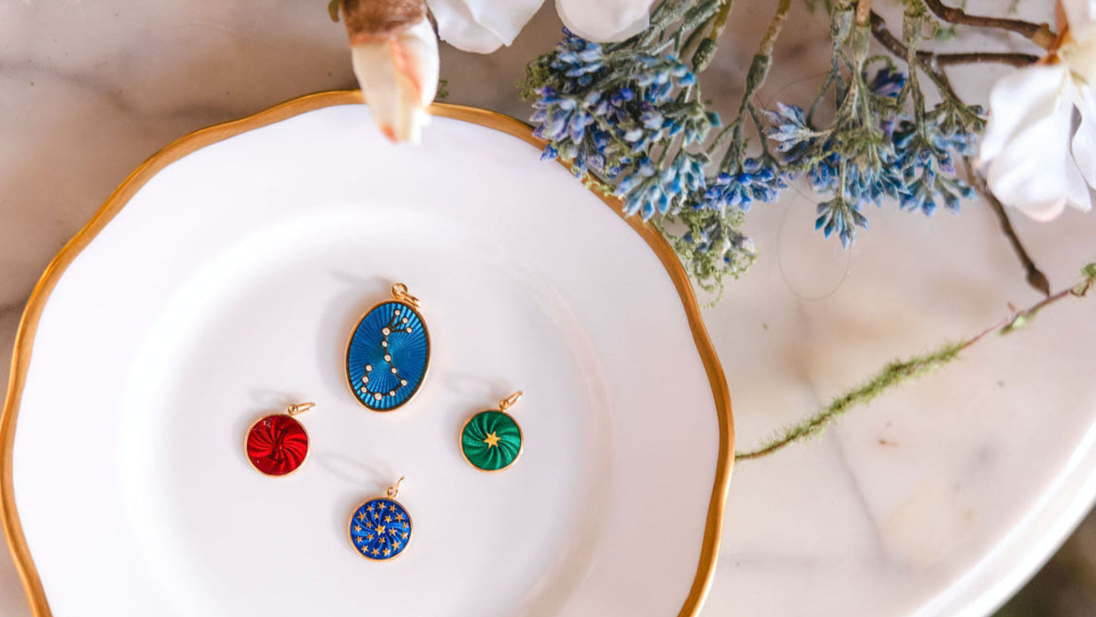 Colorful enamel guilloche pendants on a white dish with gold rim, surrounded by flowers.