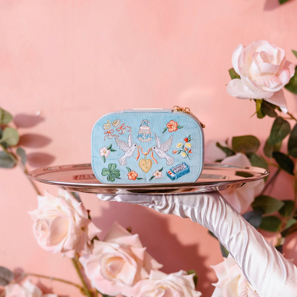 A light blue embroidered travel jewelry box with a gold zipper, held on a silver tray by a white-gloved hand against a pink background decorated with pale pink roses.