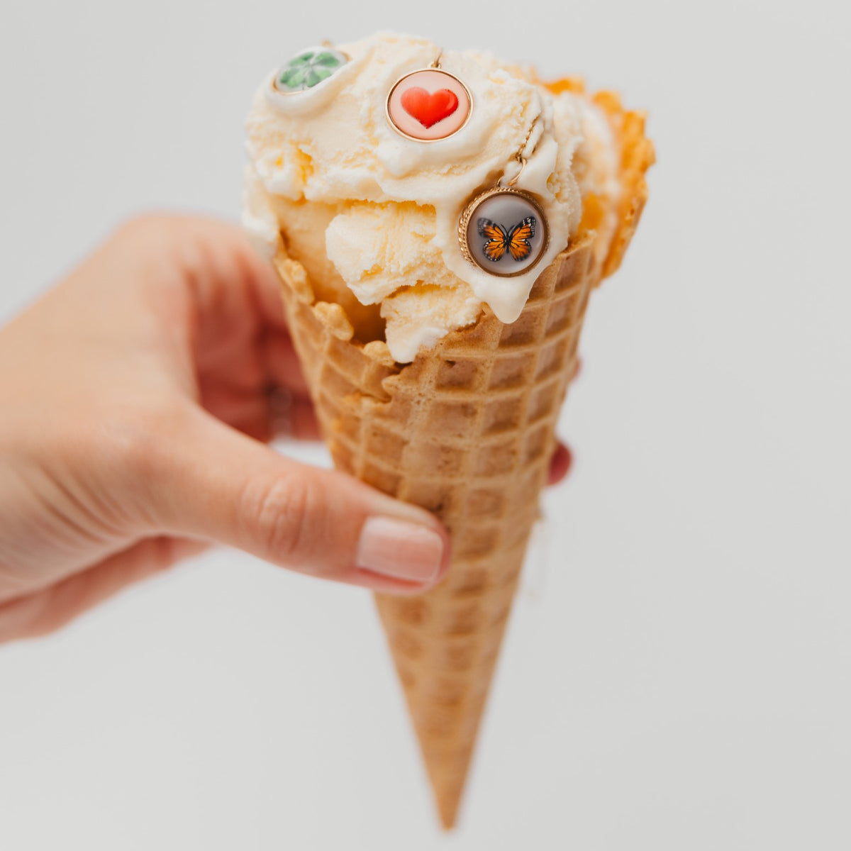 A hand holding a waffle cone filled with vanilla ice cream and topped with three decorative candy-like ornaments, against a plain light gray background.