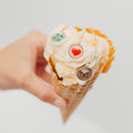 A hand holding an ice cream cone with three small pendant necklaces attached to the scoop. The necklaces have round charms: a green four-leaf clover, a red heart, and a butterfly, all on delicate chains against a plain light gray background.