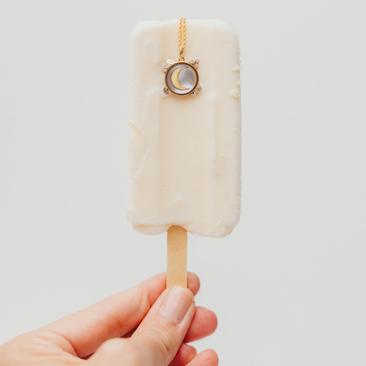 A hand holding a white popsicle-shaped pendant necklace with a gold chain and a moon charm, against a light gray background.