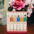 Six decorative books in a red velvet-lined box, each with a matching pendant necklace hanging from the spine. The spines are cream with gold detailing and feature colored panels in blue, green, orange, and gold.