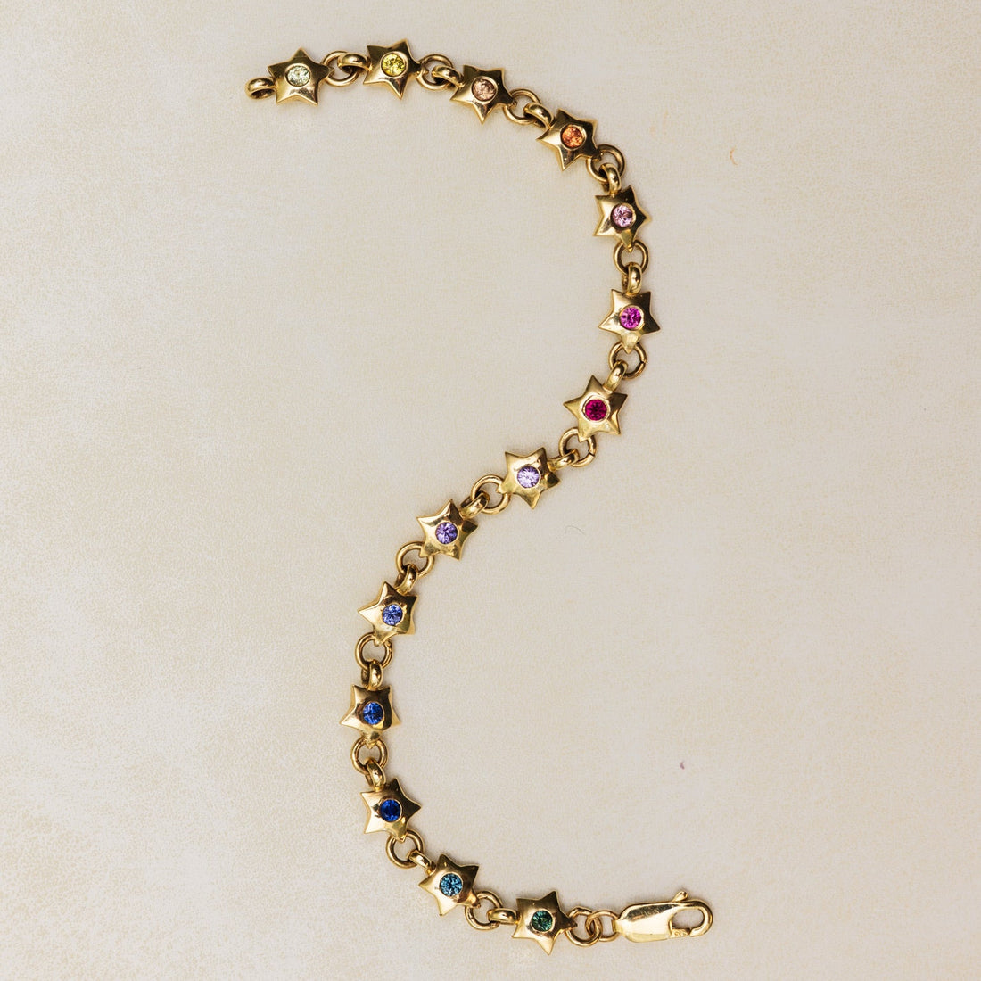 Gold bracelet with star-shaped charms and rainbow gemstones on a beige background. Rainbow Star Bracelet. Crafted in solid 14K yellow gold, each petite star is set with vibrant 0.80 carats of colored sapphires. Jewelry Dallas.