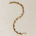 A gold star-link chain bracelet with small colorful gemstone accents, laid out in an S-shape on a light beige background.