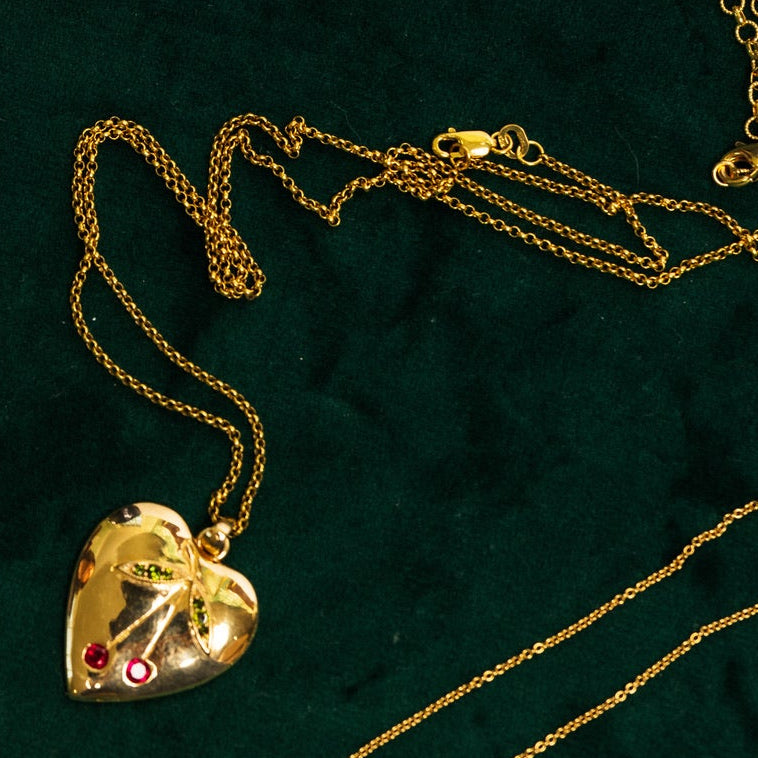 A collection of gold jewelry, including a Gold heart-shaped pendant with natural gemstones in the shape of a cherry, arranged on a dark green velvet jewelry tray.