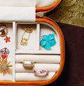 A jewelry box lined with white fabric holds several pieces of jewelry. In the upper-right compartment, the featured blue flower pendant has five bright turquoise-blue petals with finely ridged, sculpted surfaces. A small round greenish stone or bead is set at the center, surrounded by a thin metallic border. The pendant rests against the white padded lining beside gold jewelry and rings.