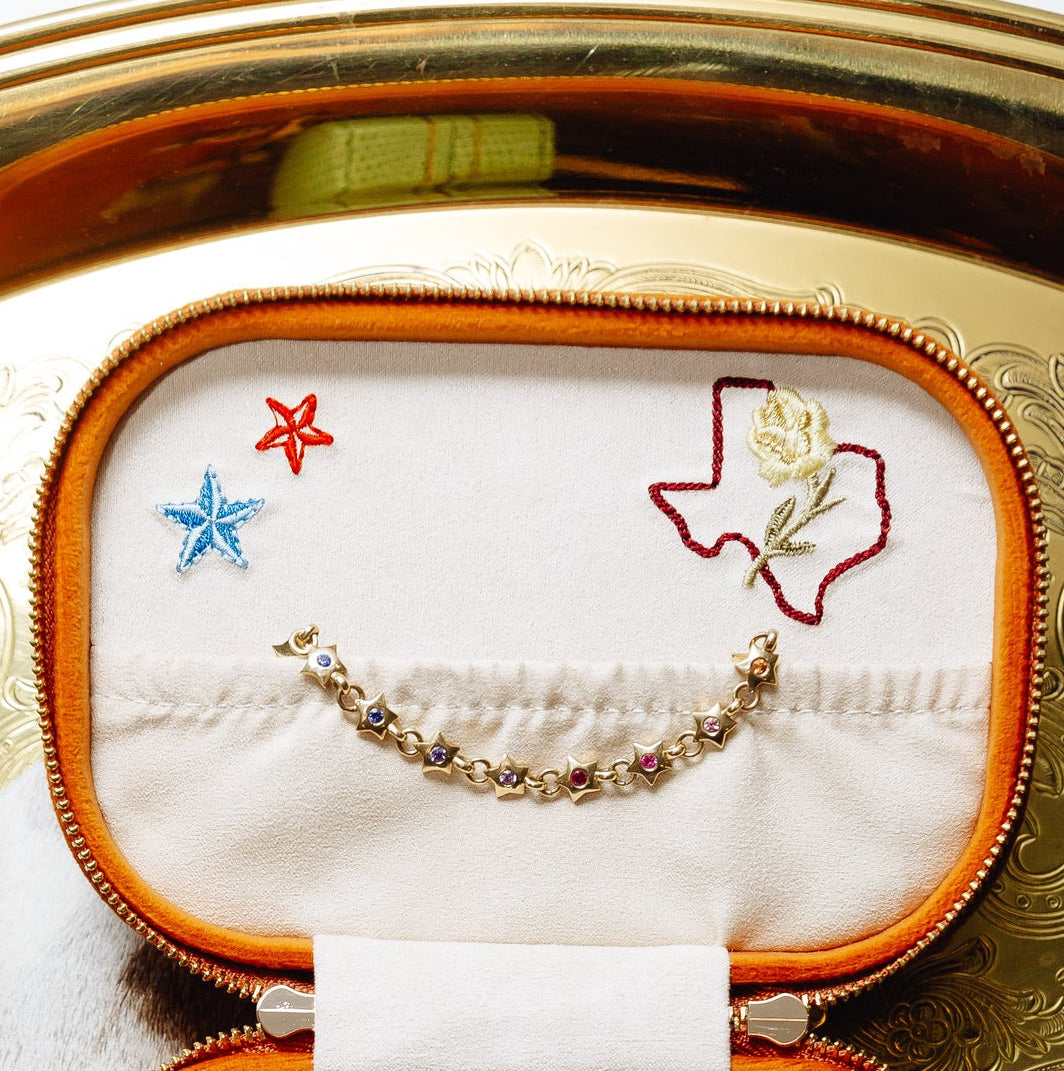 An open, ornate jewelry box displays a delicate gold-toned bracelet against white satin. The bracelet forms a shallow curve and features small star-shaped settings with blue, purple, and pink gemstones, along with linked circular elements. The interior lid is embroidered with a blue star, a red star, and a red outline of Texas containing a pale yellow flower and green stem. The box has a rounded rectangular shape with warm brown edging, beaded gold trim, and an embossed metallic-gold exterior visible around
