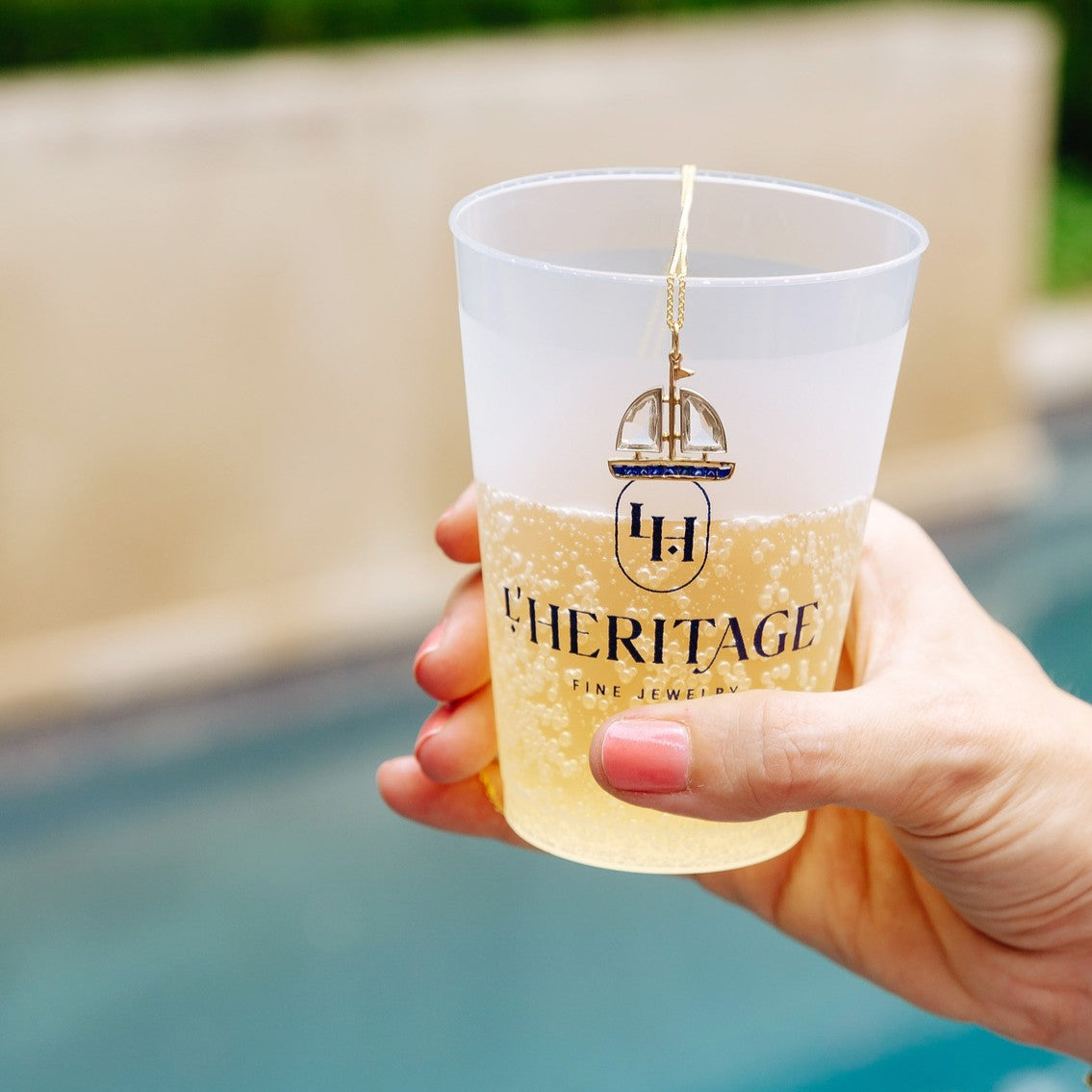 A hand holds a translucent plastic cup with a gold and navy blue 'L'Heritage Fine Jewelry' logo, filled with a bubbly pale yellow drink, against a blurred outdoor background of a pool and beige wall.