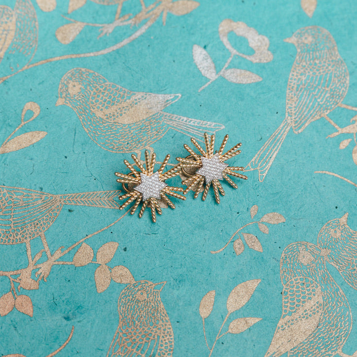 A pair of gold starburst stud earrings with white textured centers, displayed on a turquoise background with a gold bird and leaf pattern.