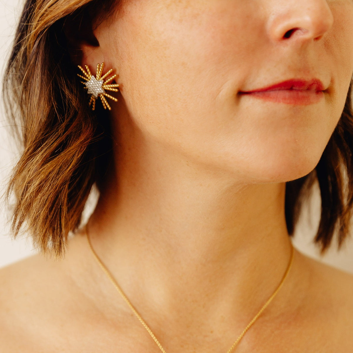 A close-up photo of a woman wearing a gold starburst stud earring with diamonds, against a soft beige background.