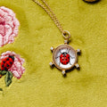 Gold ladybug pendant necklace on a textured olive green fabric background with embroidered flowers.