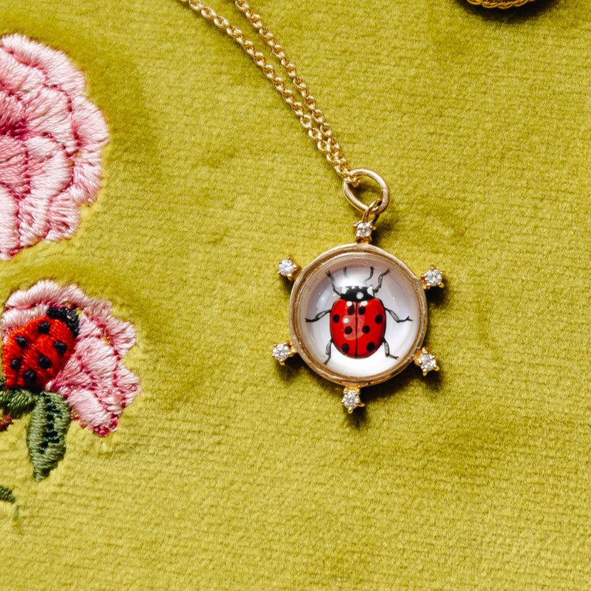 Gold ladybug pendant necklace on a textured olive green fabric background with embroidered flowers.