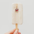 A hand holding a mini popsicle-shaped charm on a wooden stick. The charm is off-white with a small gold chain and a central ladybug charm in a circular frame.