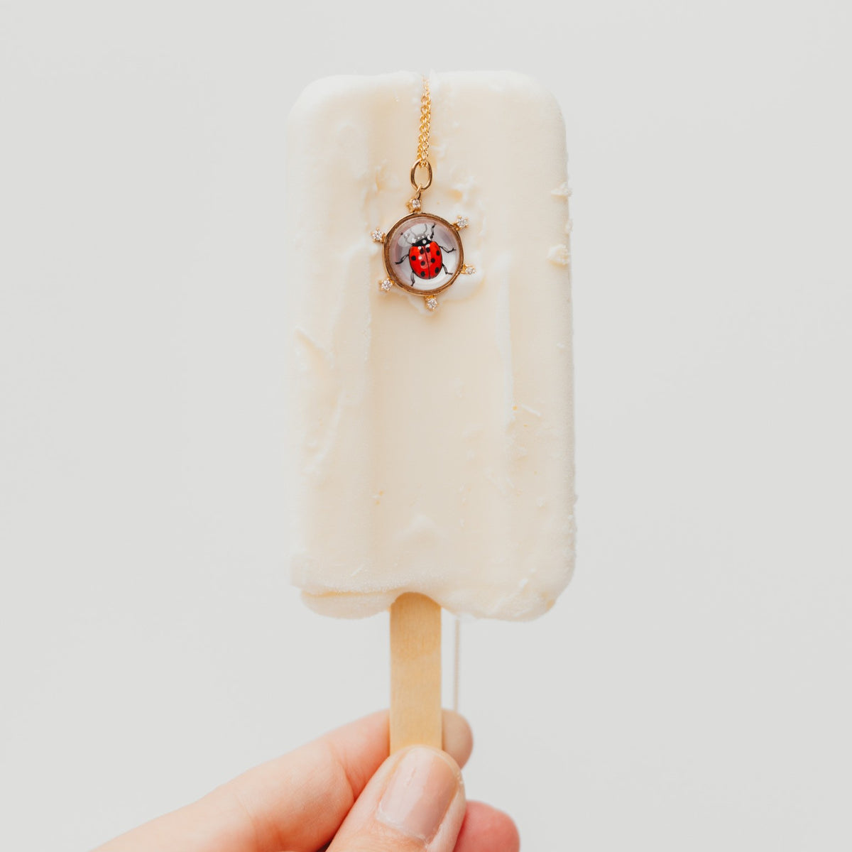 A hand holding a mini popsicle-shaped charm on a wooden stick. The charm is off-white with a small gold chain and a central ladybug charm in a circular frame.