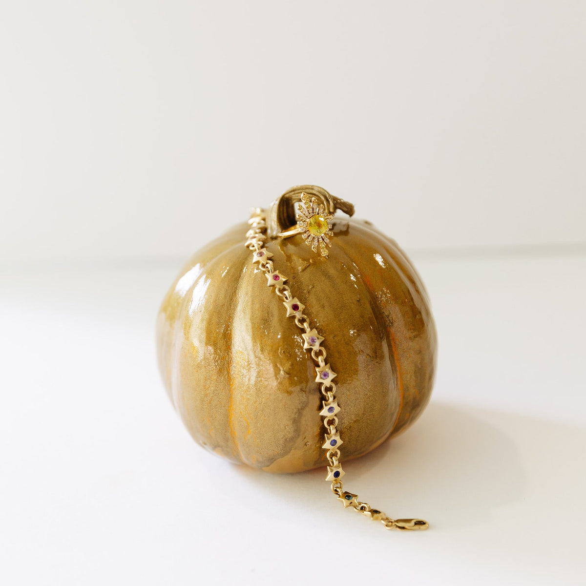 A small, round, pumpkin-shaped ceramic object with deep vertical ridges sits on a white surface against a pale background. Its glossy glaze is mottled in metallic gold, tan, and brown tones, with lighter highlights along the ridges. A delicate gold-colored chain with repeating small star- or flower-shaped links and colored stones drapes from the top of the object down its front and extends onto the surface. At the top is a gold-toned decorative clasp or pendant featuring a circular, sunburst-like design. A 